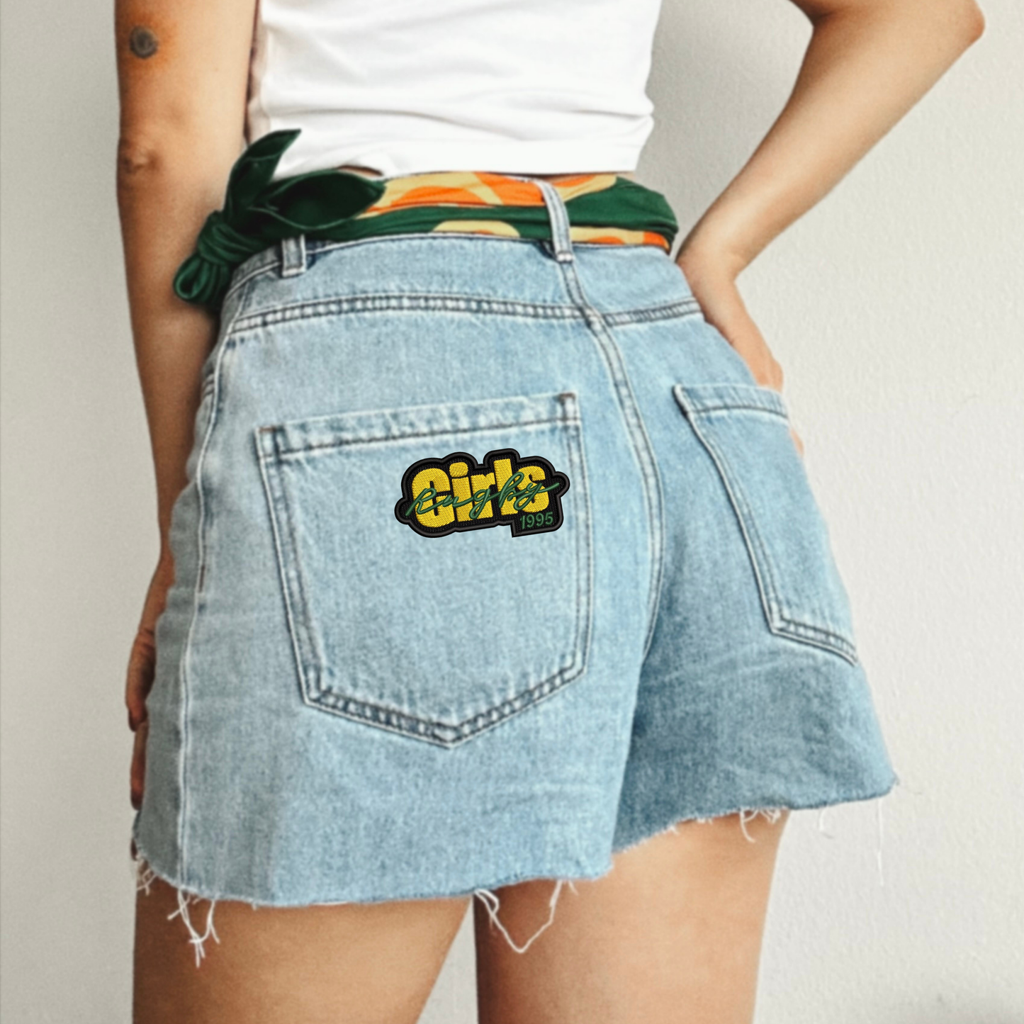 Girls Rugby 1995' embroidered patch on the back pocket of light-wash denim shorts, worn with a green and orange 1995 silk scarf tied at the waist