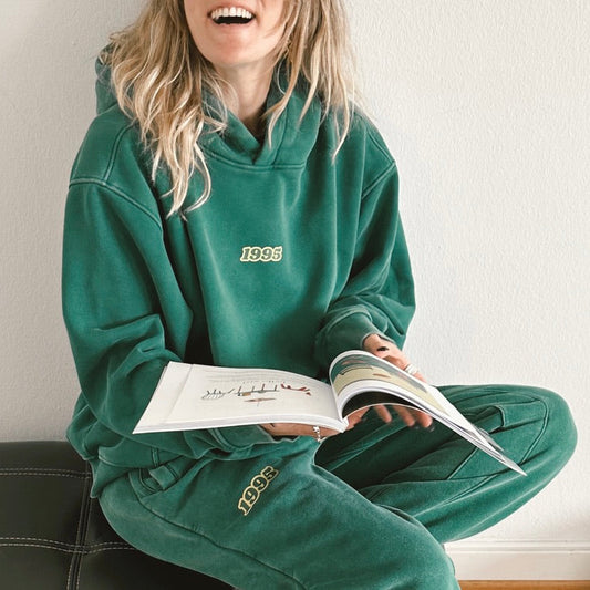 A woman wearing a matching green hoodie and sweatpants lounge set, sitting on a black leather couch and holding an open magazine. The hoodie features the year '1995' printed in gold on the chest, and the matching sweatpants have it printed on the thigh
